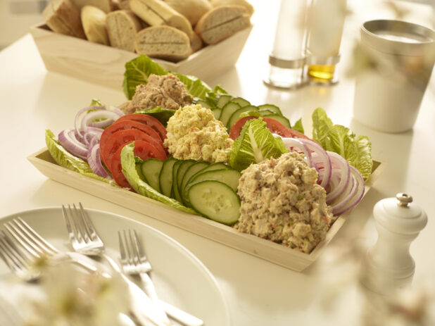 Egg salad, tuna salad and chicken salad in mounds with sliced cucumber, sliced tomatoes and sliced red onions in a wood catering box with a wood box of bread in the background with table accessories