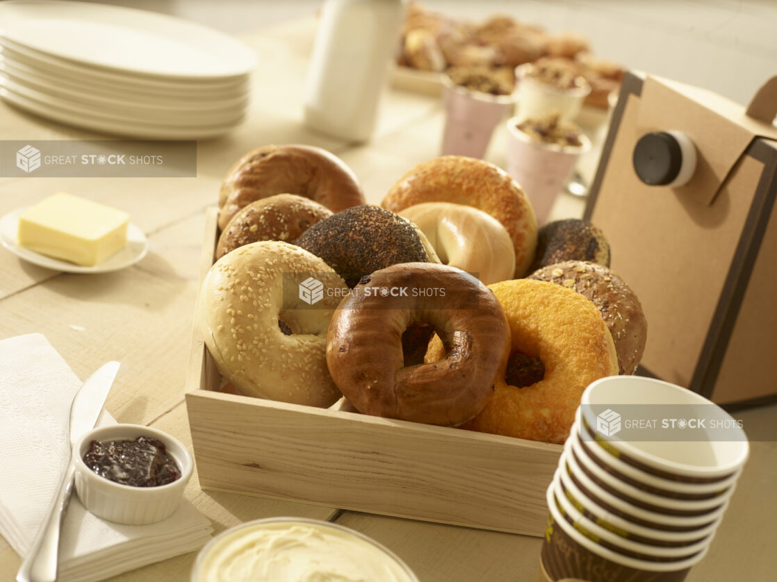 Assorted whole, uncut bagels in a wood catering box with butter, jam, cream cheese, disposable coffee cups and coffee dispenser on the side with other breakfast items in the background