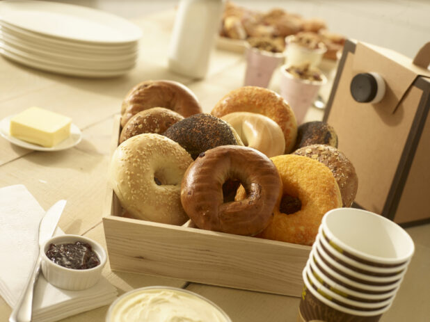 Assorted whole, uncut bagels in a wood catering box with butter, jam, cream cheese, disposable coffee cups and coffee dispenser on the side with other breakfast items in the background