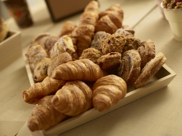 Assorted pastries on a square wood catering tray with croissants, danishes, muffins and oatmeal bites on a wooden background in a close up view