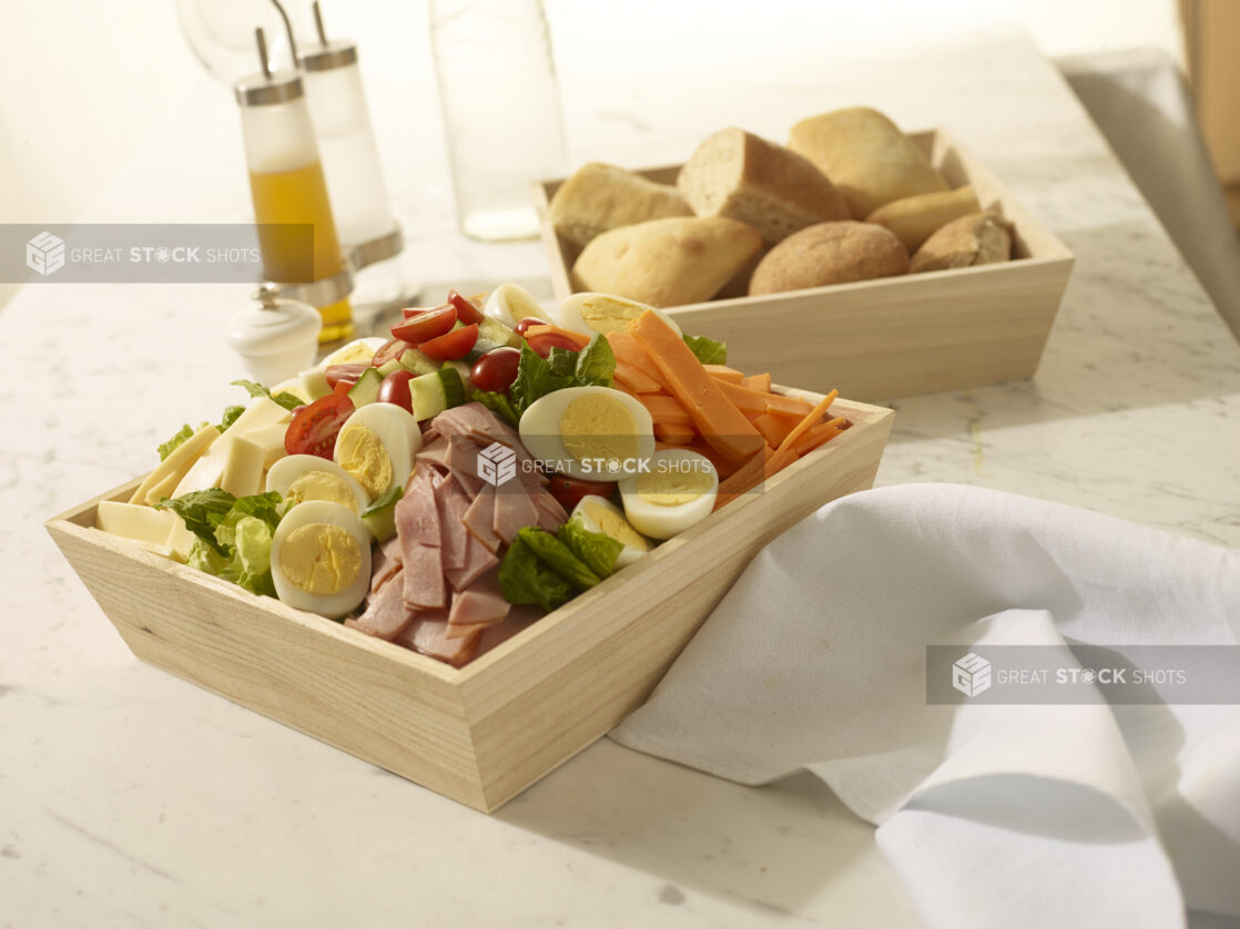 Chef salad with ham, cheese, boiled eggs, carrots, cucumber and tomatoes in a wood catering box with a wood catering box of bread rolls in the background