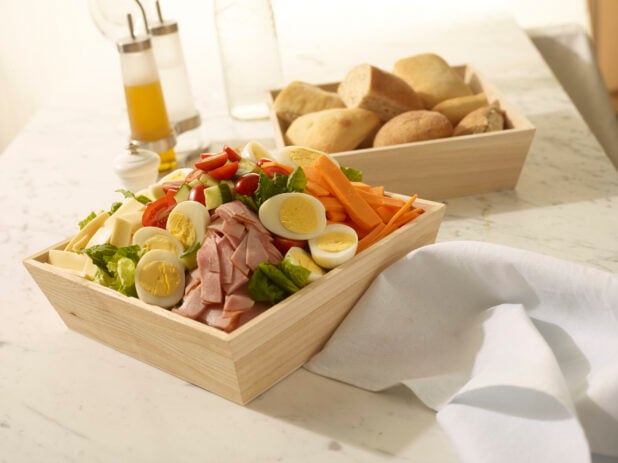 Chef salad with ham, cheese, boiled eggs, carrots, cucumber and tomatoes in a wood catering box with a wood catering box of bread rolls in the background