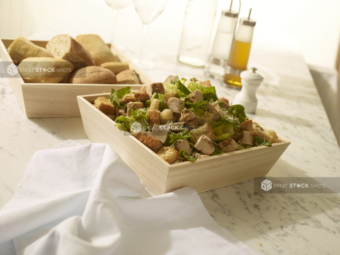 Chicken Caesar salad with croutons and parmesan cheese in a wood catering box with bread in a wood catering box in the background on a white marble background