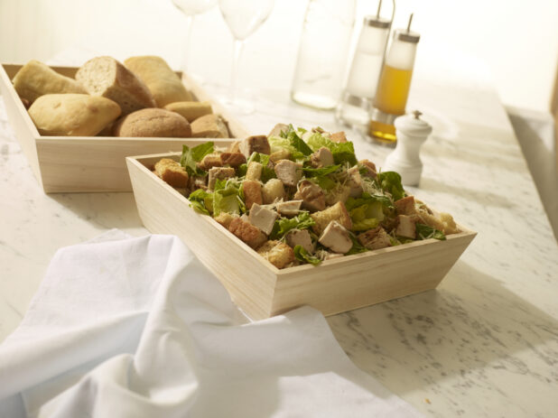 Chicken Caesar salad with croutons and parmesan cheese in a wood catering box with bread in a wood catering box in the background on a white marble background