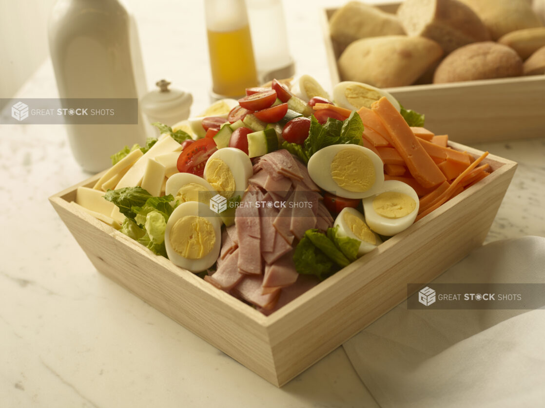 Chef salad with ham, cheese, boiled eggs, carrots, cucumber and tomatoes in a wood catering box with a wood catering box of bread rolls in the background