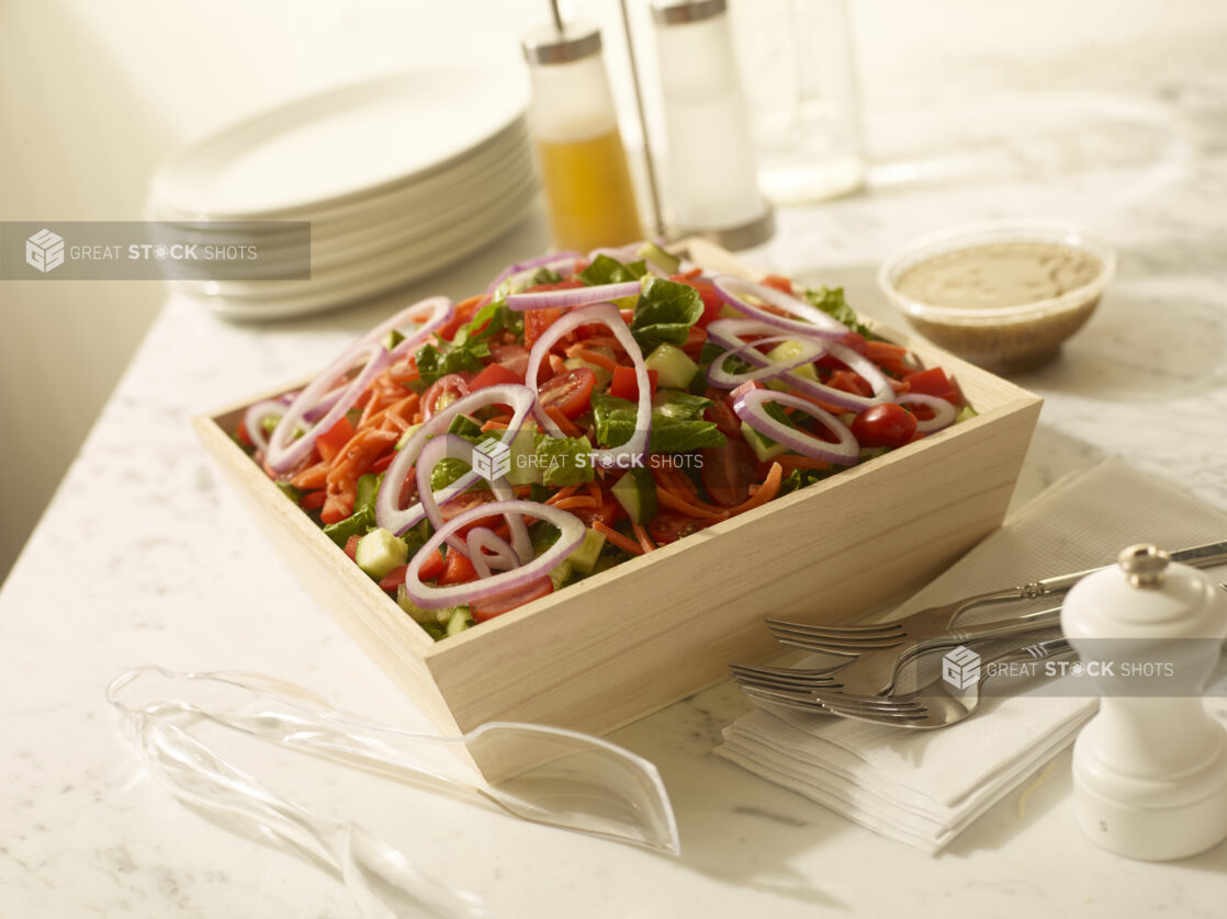 Deep wood catering tray of garden salad with a side dressing in a glass dish and plastic tongs on the side on a white marble countertop