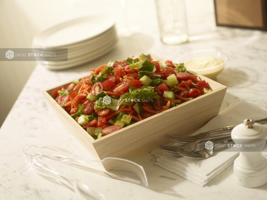 Garden salad with cucumber, tomato and shredded carrots in a wood catering box with forks, napkins, side plates, salad dressing and serving tongs on a white a marble background