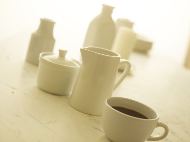 Cup of coffee in a white cup with a white cream pitcher and white sugar bowl in the background, close-up