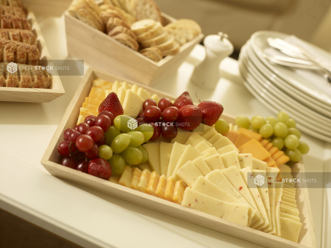 Cheese platter on a wood catering tray with assorted cheeses, grapes and strawberries with other party platters surrounding, tilted angle