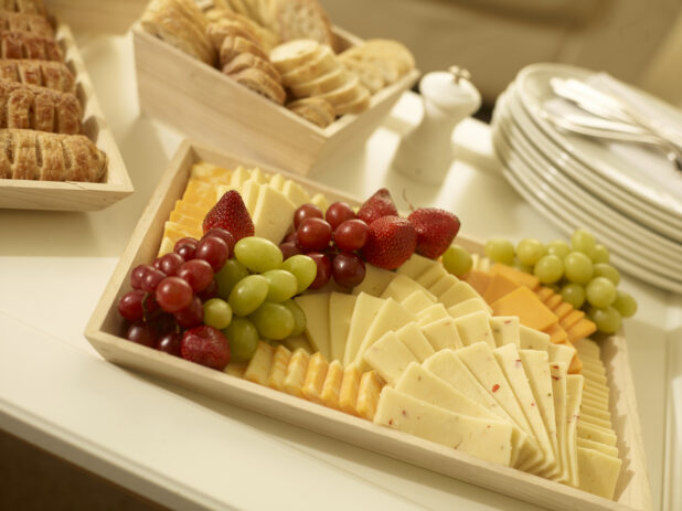 Cheese platter on a wood catering tray with assorted cheeses, grapes and strawberries with other party platters surrounding, tilted angle