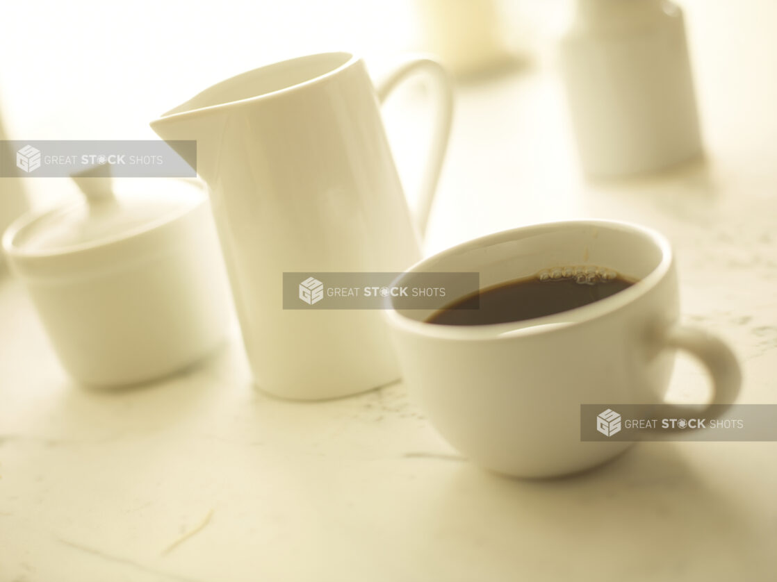 Cup of coffee in a white cup with a white cream pitcher and white sugar bowl in the background