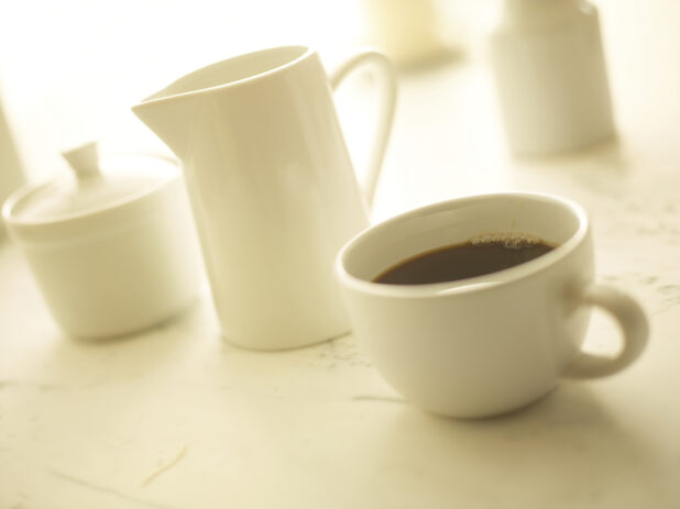 Cup of coffee in a white cup with a white cream pitcher and white sugar bowl in the background