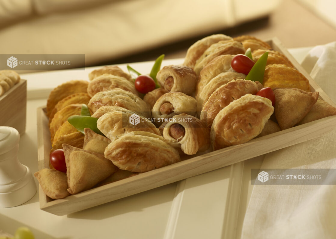 Appetizer platter with mini sausage rolls, mini samosas, mini jamaican patties, mini cornish pasties on a square wood catering tray