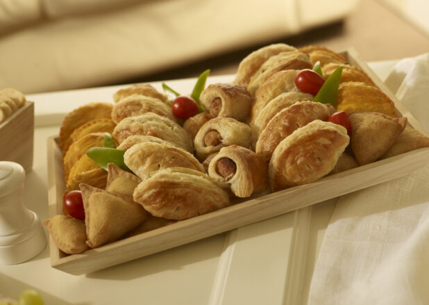 Appetizer platter with mini sausage rolls, mini samosas, mini jamaican patties, mini cornish pasties on a square wood catering tray