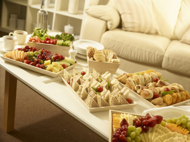 Party scene with veggies and dip, cheese tray, party sandwiches, appetizer platter, fruit tray and bread box on a coffee table with white side plates