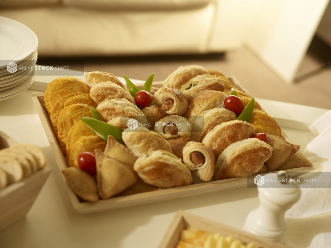 Appetizer platter with mini sausage rolls, mini samosas, mini jamaican patties, and mini cornish pasties on a square wood catering tray in a party setting