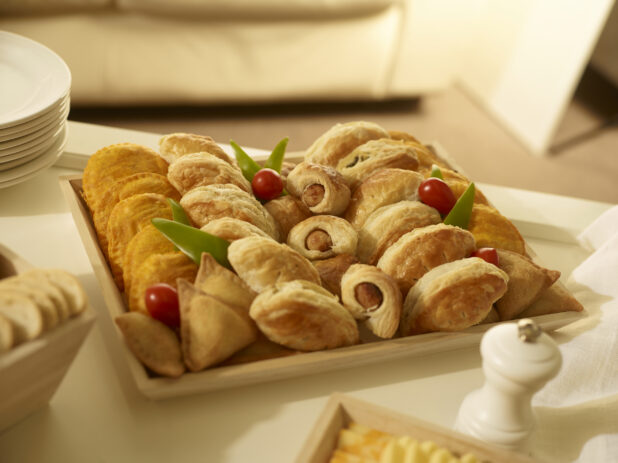 Appetizer platter with mini sausage rolls, mini samosas, mini jamaican patties, and mini cornish pasties on a square wood catering tray in a party setting