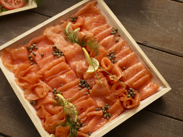 Smoked salmon in a square wood catering box with sliced lemons, capers and dill garnish on a wooden background