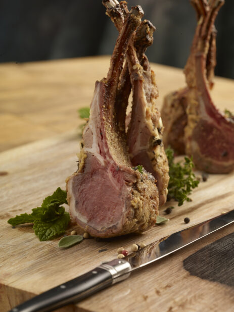 Roast rack of lamb, sliced, standing up on a wooden board with mint and peppercorn garnish with a carving knife on the side