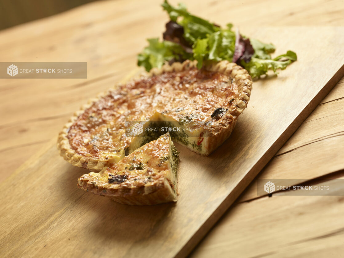 Vegetarian quiche, one slice cut out with mixed greens in the background all on a wooden board and background