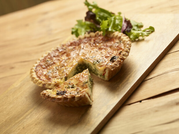 Vegetarian quiche, one slice cut out with mixed greens in the background all on a wooden board and background