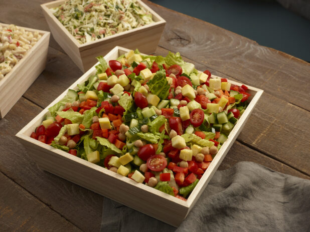Chopped vegetable salad, coleslaw and macaroni salad in wood catering trays on a wood table