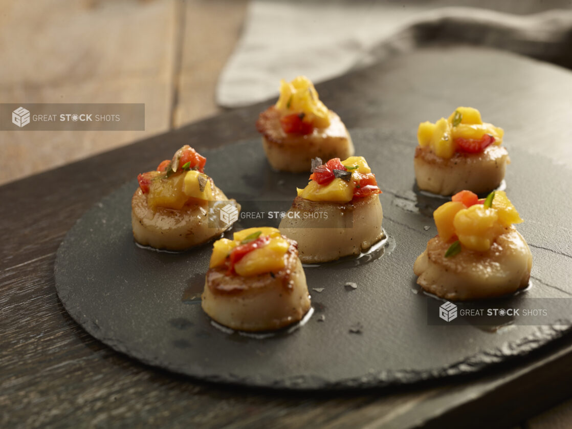 Pan seared scallops topped with mango salsa on a round slate platter on a wooden background