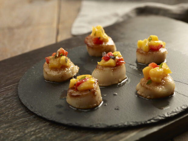 Pan seared scallops topped with mango salsa on a round slate platter on a wooden background