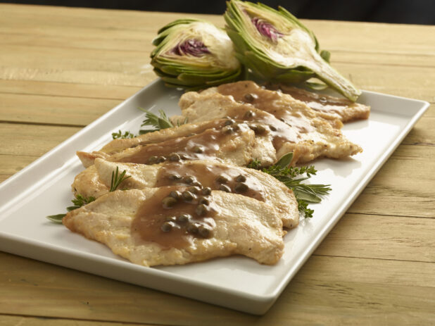 Chicken scallopine with gravy and capers garnished with fresh herbs and a fresh artichoke cut in half on a rectangular white plate on a wooden background