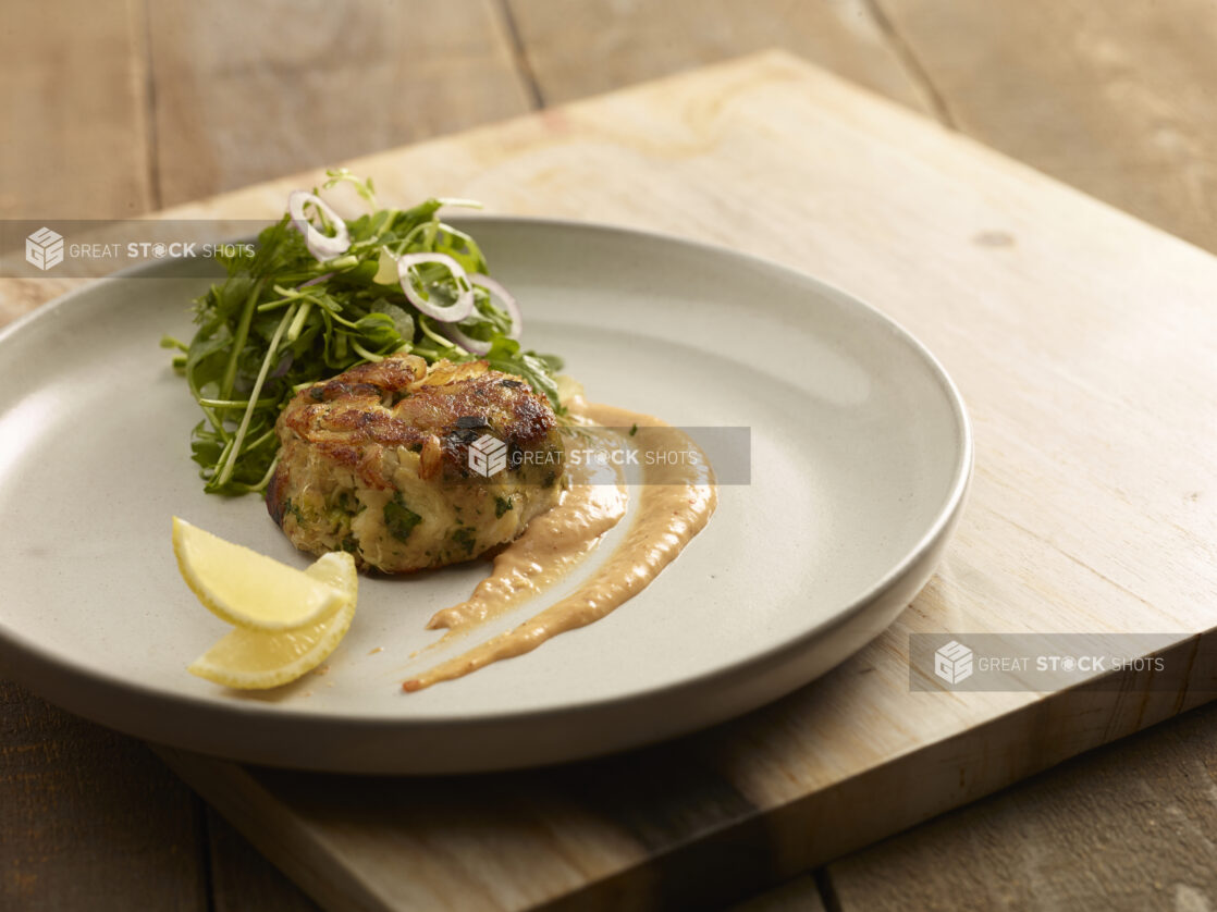 Crab cake with a side green salad on a plate with a sauce smear and two lemon wedges on a wooden board