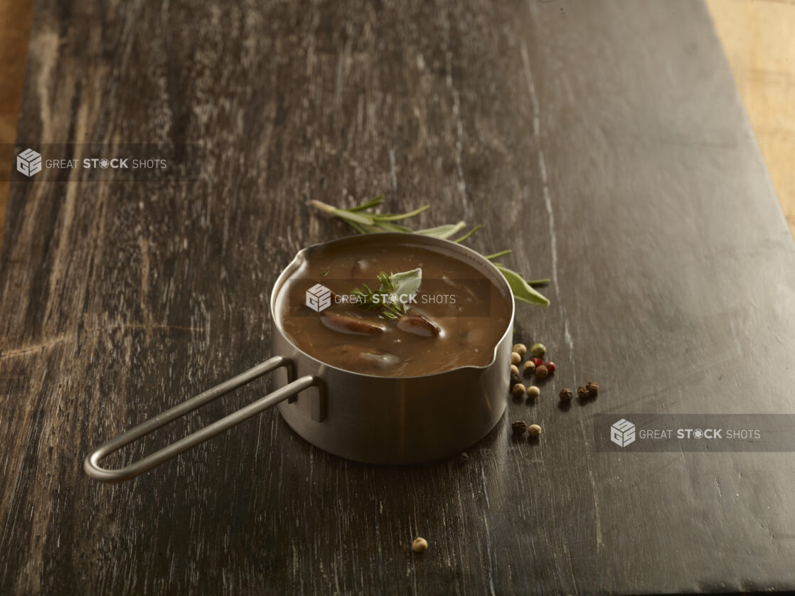 Gravy in a metal pouring pot with a sprig of rosemary and peppercorns on the side on a dark wooden board