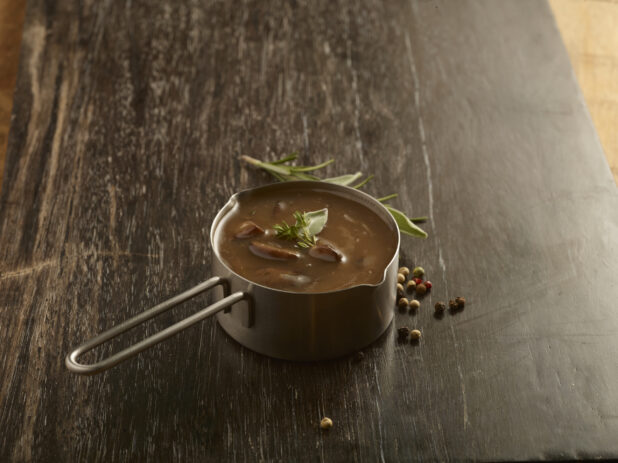 Gravy in a metal pouring pot with a sprig of rosemary and peppercorns on the side on a dark wooden board