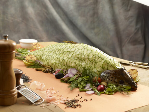 Whole poached fish with cucumber slices covering the fish with fresh herbs, vegetables, grilled lemon surrounding, salt shaker, pepper mill with pink salt and peppercorns on the side