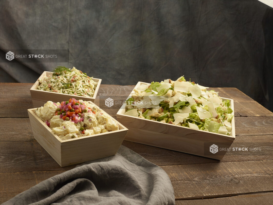 Caesar salad, potato salad and coleslaw in large wood catering bowls on a wood table