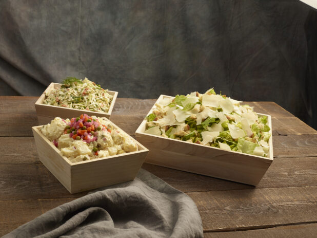 Caesar salad, potato salad and coleslaw in large wood catering bowls on a wood table
