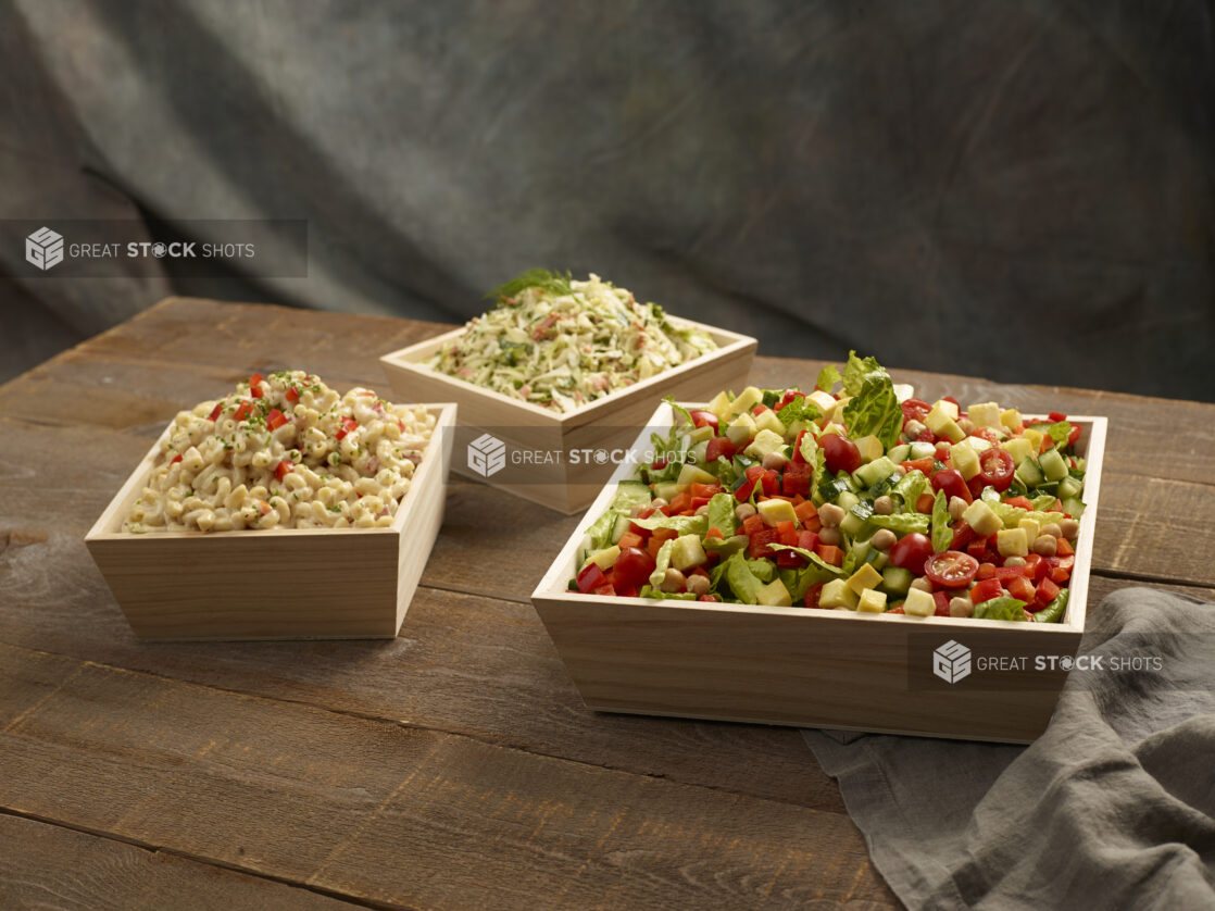 Green salad with fresh vegetables, macaroni salad and coleslaw in wood catering boxes on a wooden background,