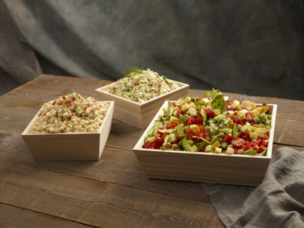 Green salad with fresh vegetables, macaroni salad and coleslaw in wood catering boxes on a wooden background,