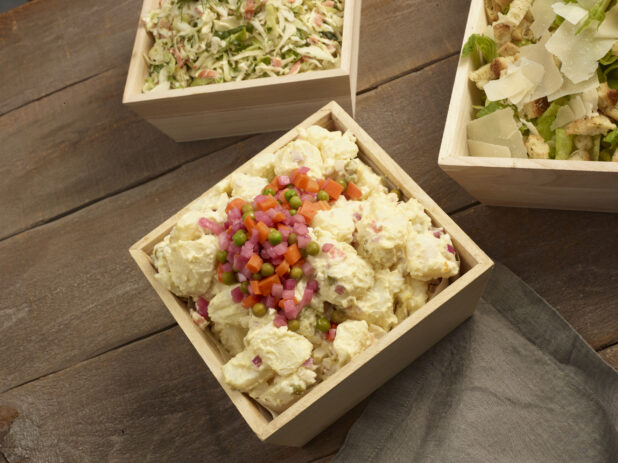 Potato salad, coleslaw and Caesar salad all in square wooden catering boxes on a wooden background