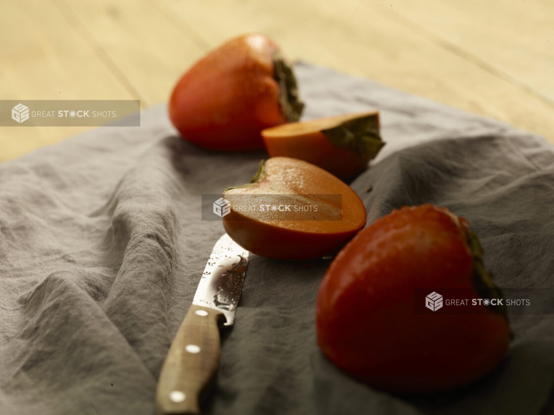 Whole and sliced fresh persimmons on a grey cloth background with a paring knife on a wooden background, close up view