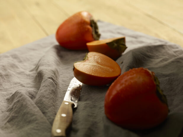 Whole and sliced fresh persimmons on a grey cloth background with a paring knife on a wooden background, close up view