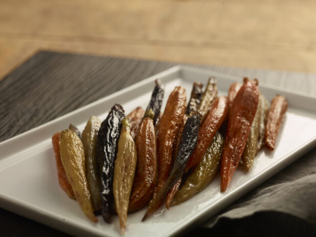 Roasted heirloom carrots on a white rectangular plate on a dark wooden board, on a wooden background