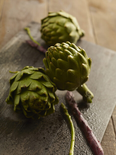 3 whole fresh artichoke with a couple of purple and green asparagus stems on a dark wooden board on a wooden background