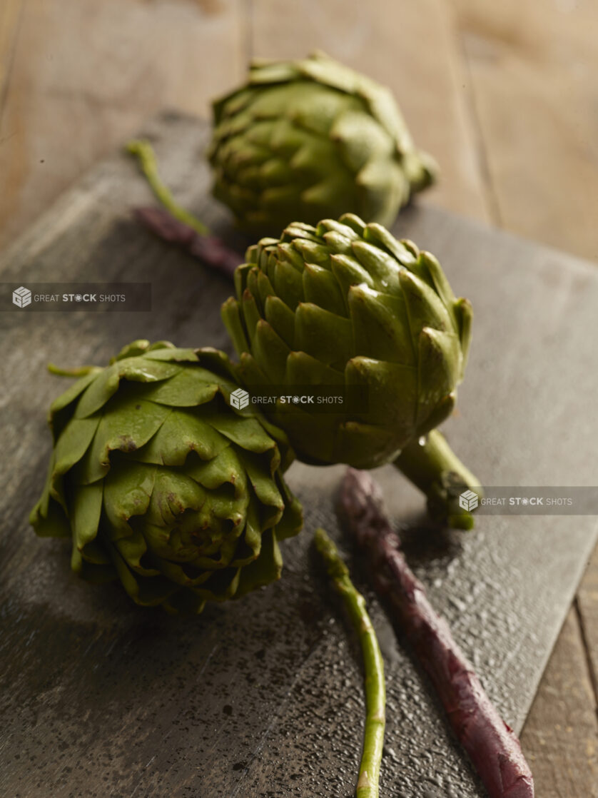 3 whole fresh artichoke with a couple of purple and green asparagus stems on a dark wooden board on a wooden background