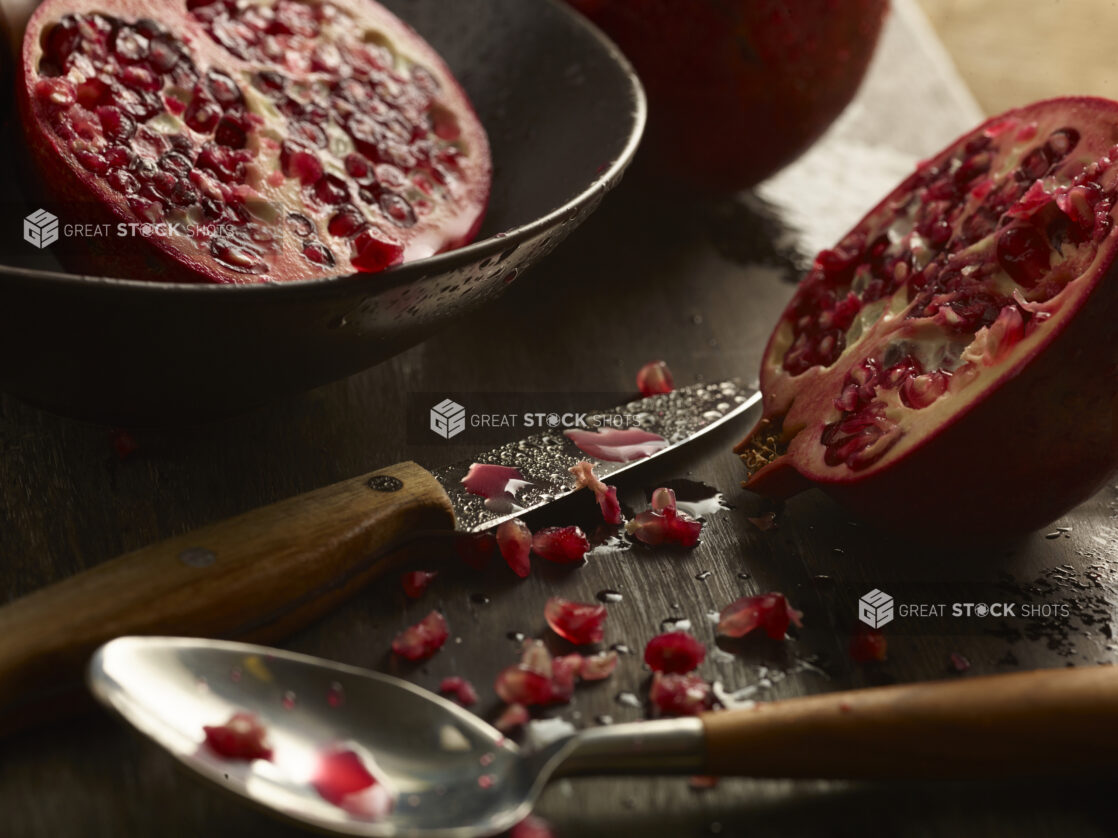 Pomegranate cut in half, half in bowl, half on slate board with knife and spoon