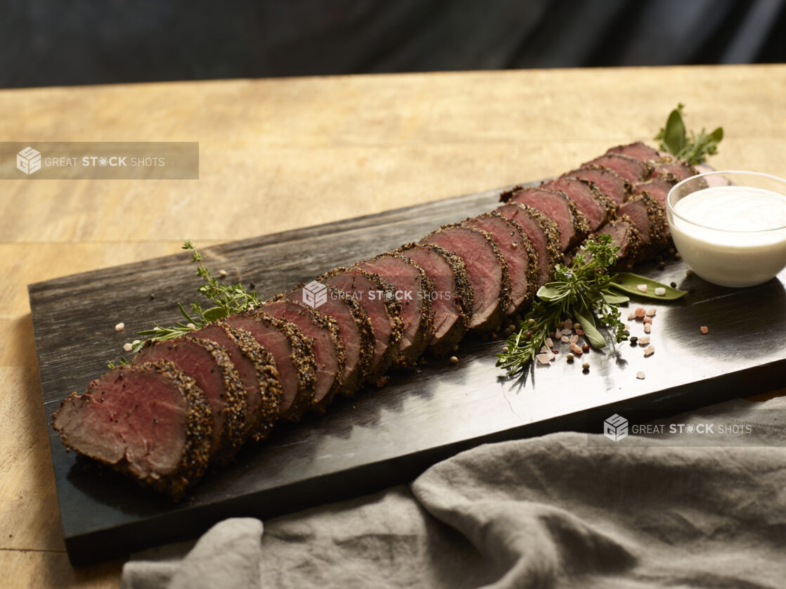 Rare beef tenderloin sliced on a dark wooden board with a glass ramekin of horseradish aioli, garnished with fresh herbs and coarse salt and pepper