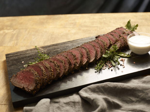 Rare beef tenderloin sliced on a dark wooden board with a glass ramekin of horseradish aioli, garnished with fresh herbs and coarse salt and pepper