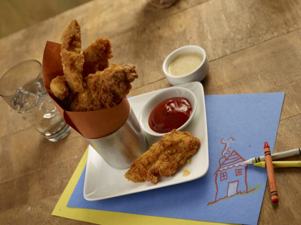 Chicken fingers in a metal cup with a rust coloured parchment paper with ketchup and dipping sauce on a square white side plate with crayons and colouring paper as placemat