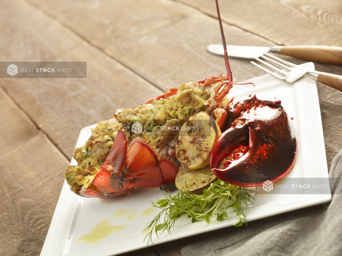 Lobster thermidor on a square white plate with grilled lemons and fresh herb garnish with a fork and knife on the side on a wooden background