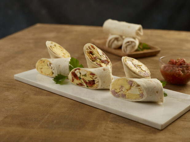 Assorted breakfast wraps cut in half on a rectangular marble board with a glass bowl of salsa on the side with whole wraps on a wood tray in the background