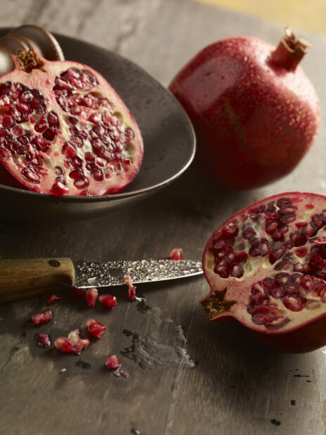 Pomegranate cut in half with one half in a bowl and the other on a wood table with seeds around the base